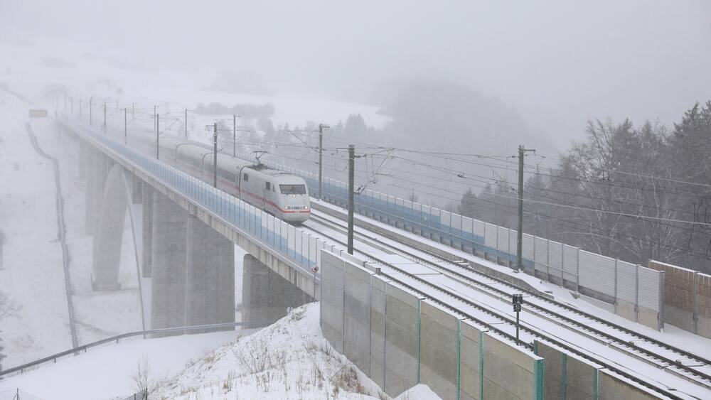 A train on a snow-covered bridge with a snowy landscape and power lines.