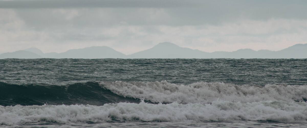 A turbulent sea with waves crashing onto the shore, a cloudy sky, and distant mountains.