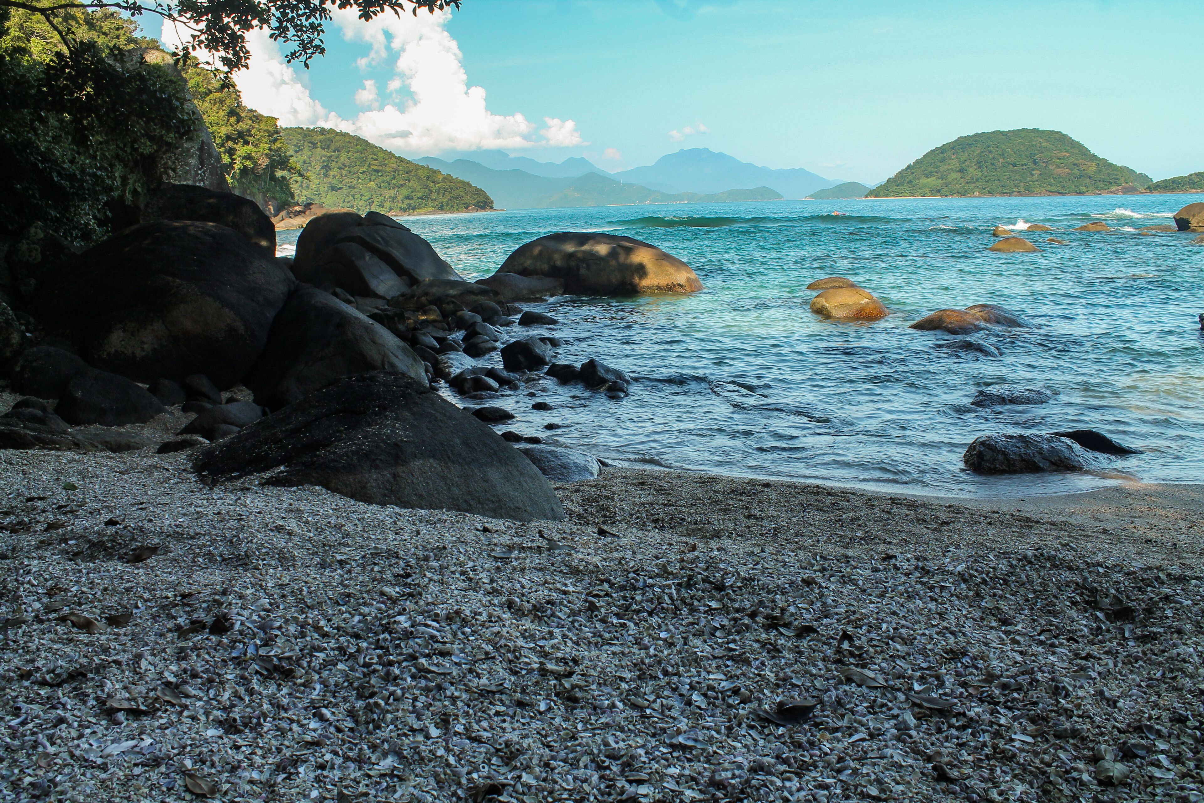 Deserted Praia das Conchas with clear waters, rocks in the background, and the lush Ilha do Prumirim on the horizon. A tranquil and scenic beach in Ubatuba, São Paulo, Brazil, on a sunny day.