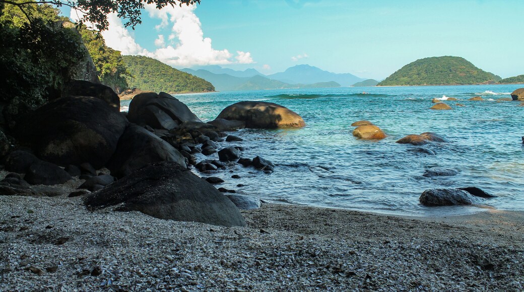 Deserted Praia das Conchas with clear waters, rocks in the background, and the lush Ilha do Prumirim on the horizon. A tranquil and scenic beach in Ubatuba, São Paulo, Brazil, on a sunny day.