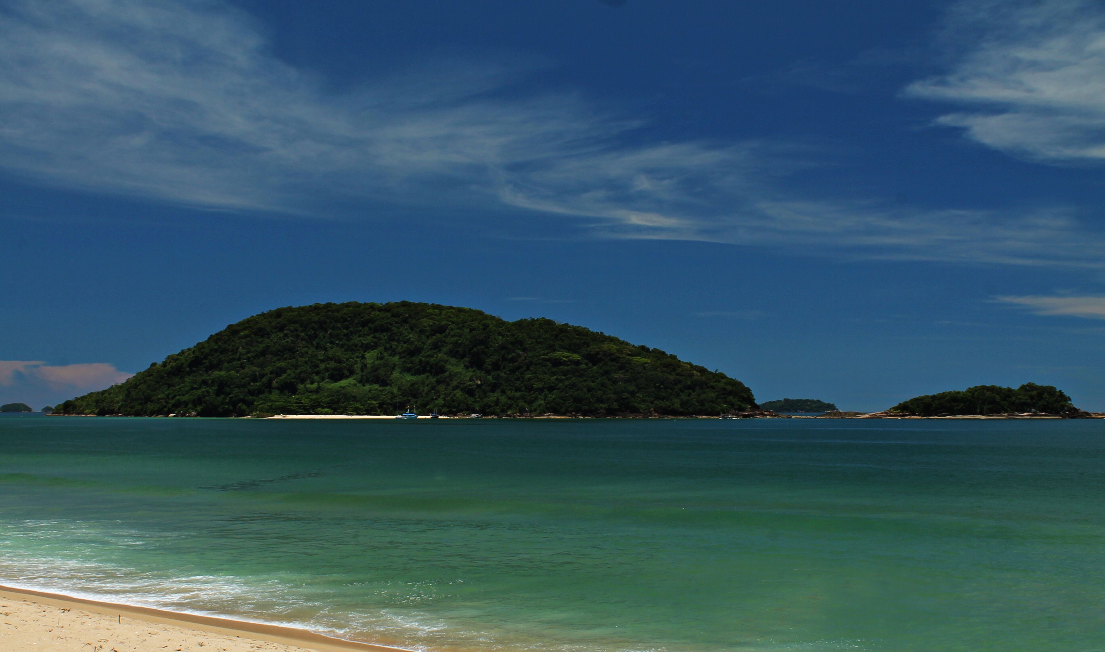 Praia do Prumirim, Ubatuba/SP, com a Ilha do Prumirim ao fundo, em dia ensolarado, águas cristalinas e montanhas cobertas por floresta tropical, destacando a natureza exuberante