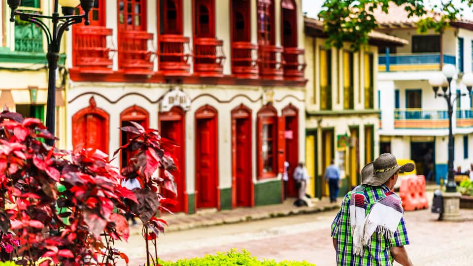 A man in a hat and colorful shirt walking past a building with red doors and balconies.
