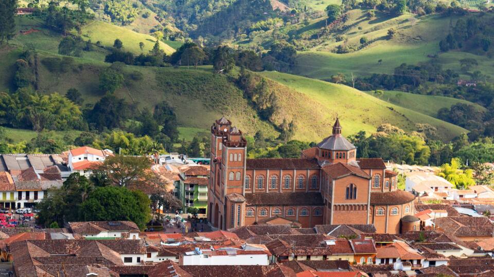 A town with terracotta-roofed buildings, a prominent church with a bell tower, and rolling green hills in the background.
