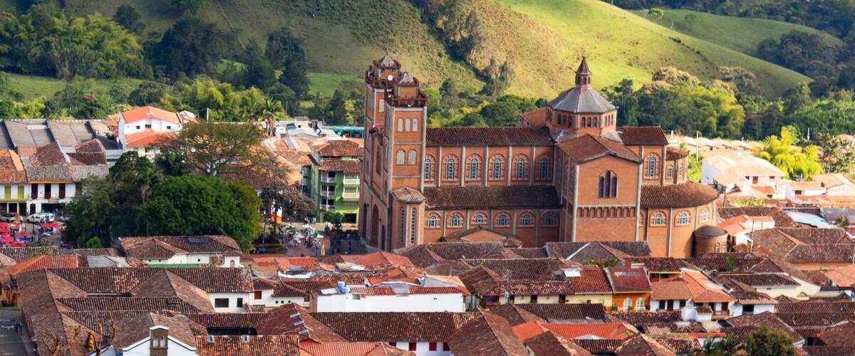 A town with terracotta-roofed buildings, a prominent church with a bell tower, and rolling green hills in the background.