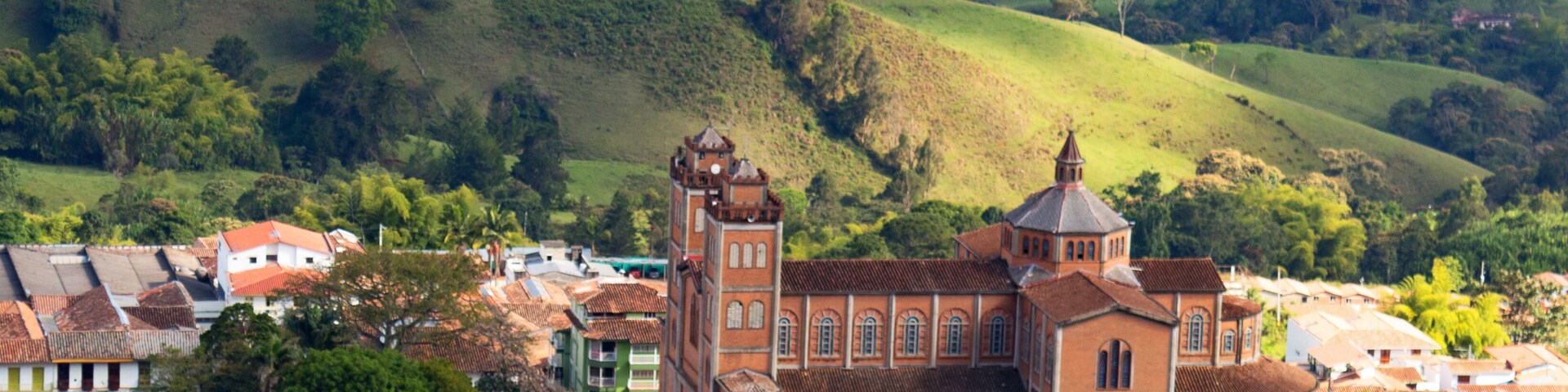 A town with terracotta-roofed buildings, a prominent church with a bell tower, and rolling green hills in the background.