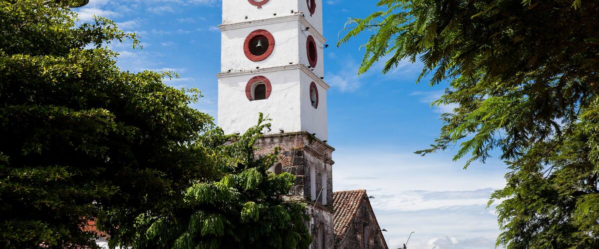 A white clock tower with red accents, surrounded by lush green trees.