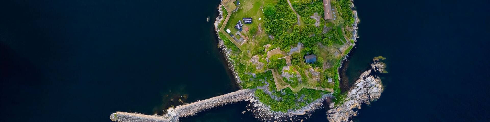An aerial view of a small island with a fortified structure, lush greenery, and a bridge connecting to the mainland.