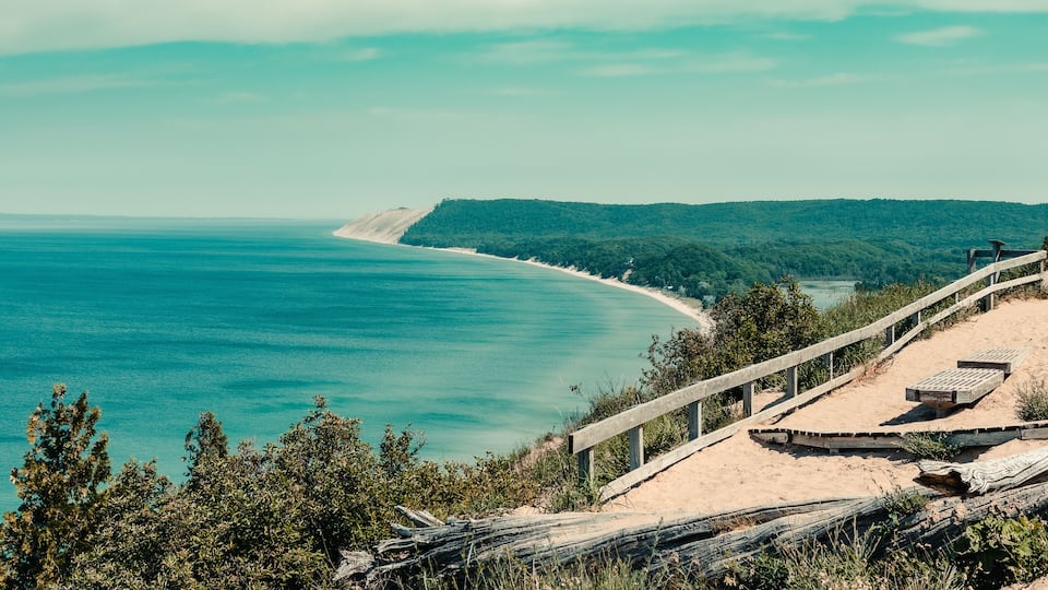 A coastal landscape with a sandy beach, a wooden walkway, and a forested hill in the background.