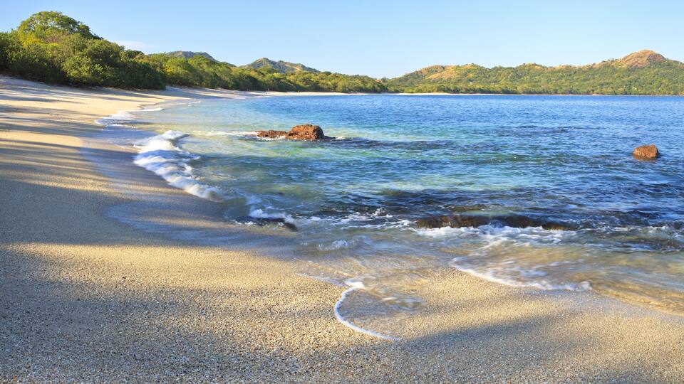 A pebbly beach with clear water, small rocks, and distant green hills.