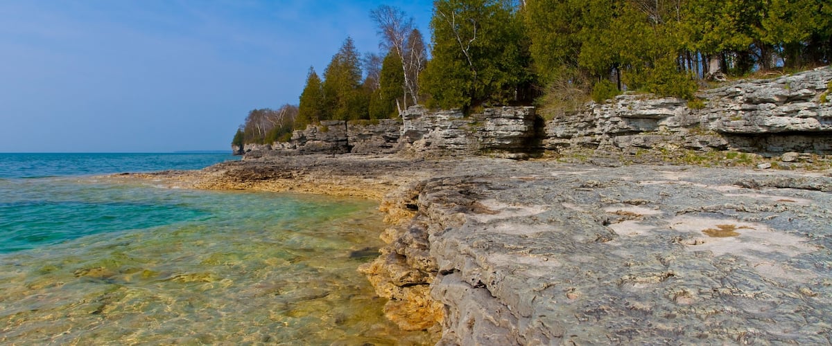 A rocky shoreline with clear turquoise water, a forested cliff, and a clear sky.