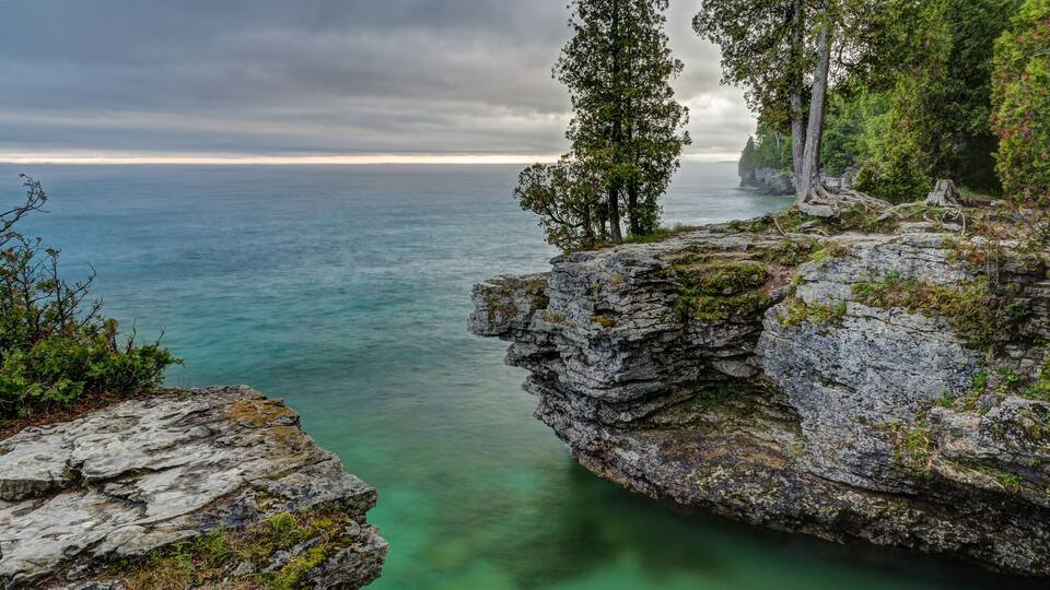A rocky coastline with clear turquoise water, a cliff with trees, and a cloudy sky.
