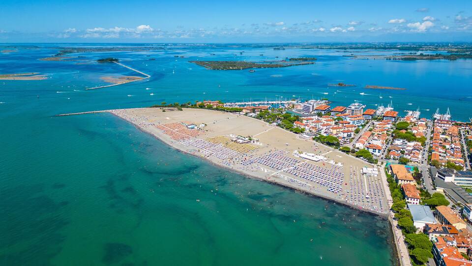An aerial view of a coastal town with a large beach, clear blue waters, and a marina with boats.