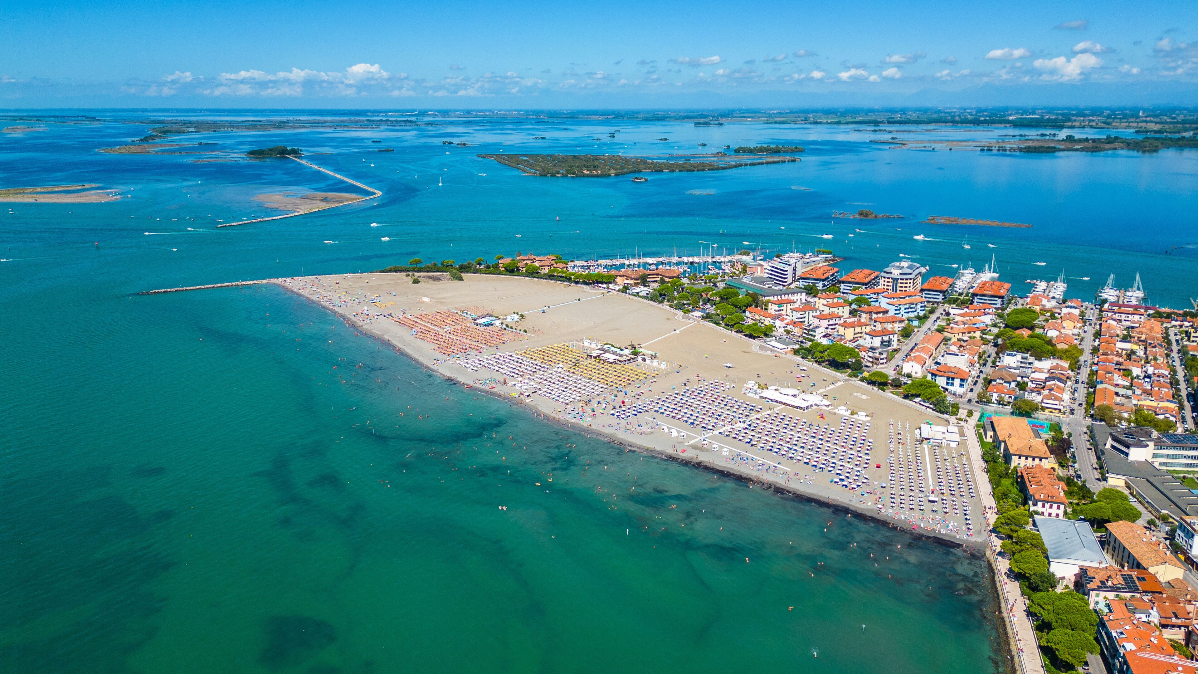 An aerial view of a coastal town with a large beach, clear blue waters, and a marina with boats.