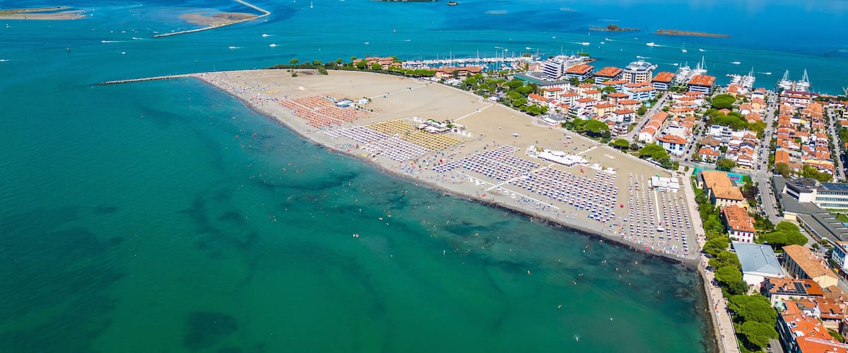An aerial view of a coastal town with a large beach, clear blue waters, and a marina with boats.