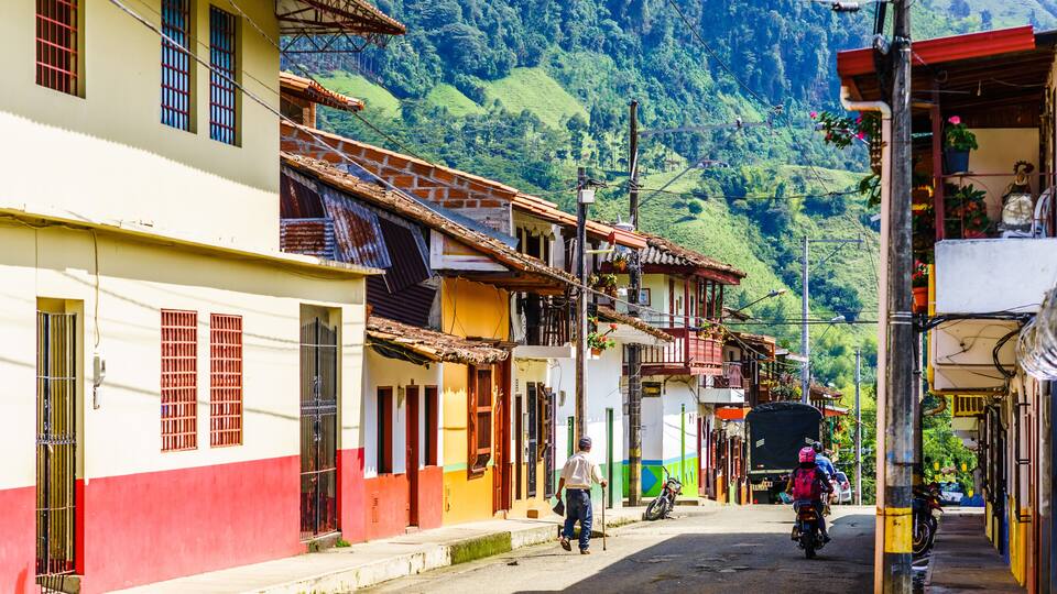 A street in a hilly town with colorful buildings, a man with a cane, and parked motorcycles.