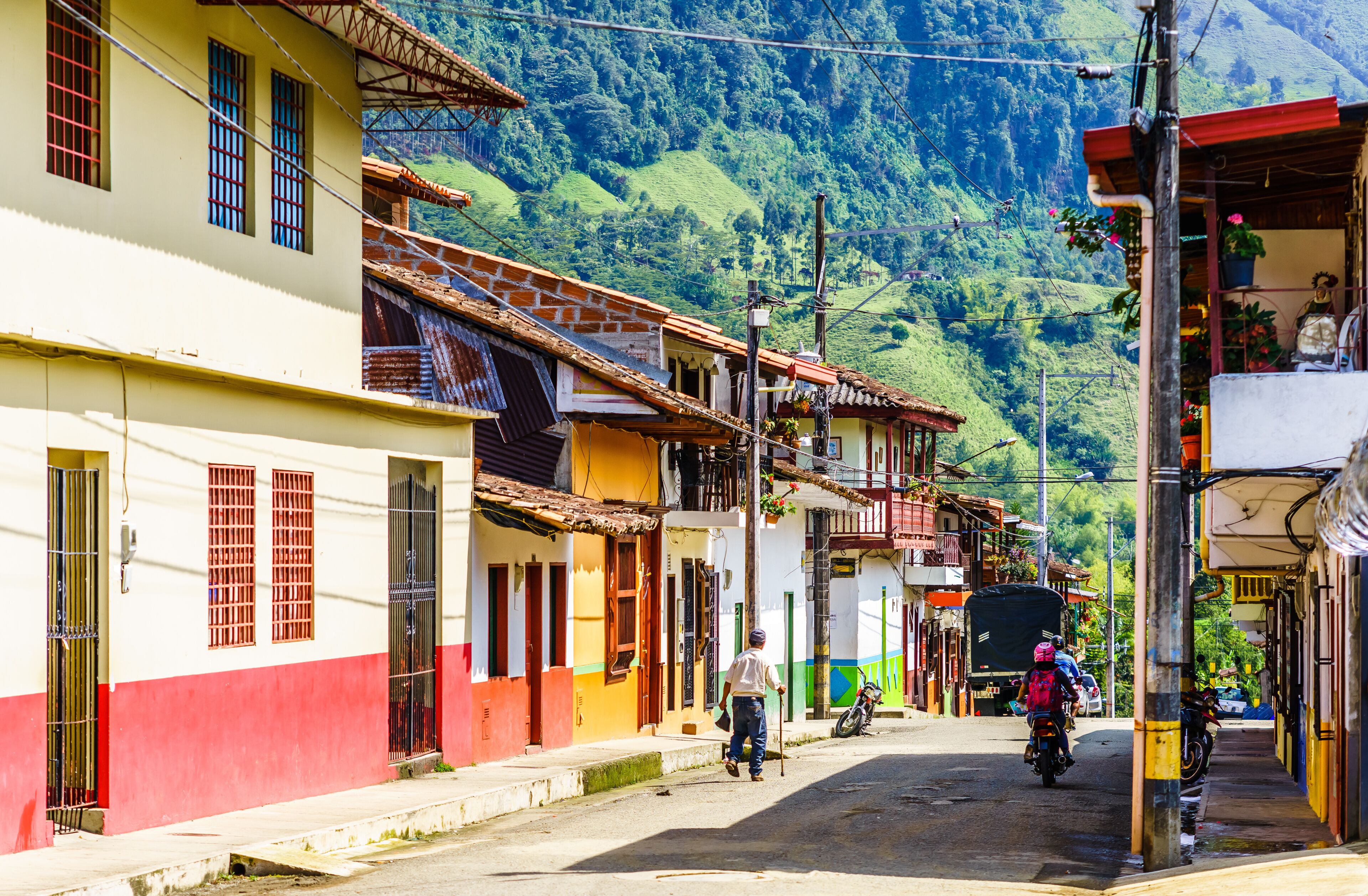 A street in a hilly town with colorful buildings, a man with a cane, and parked motorcycles.