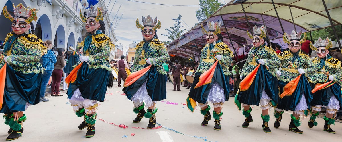 A group of performers in elaborate costumes with crowns and colorful sashes, walking in a parade.