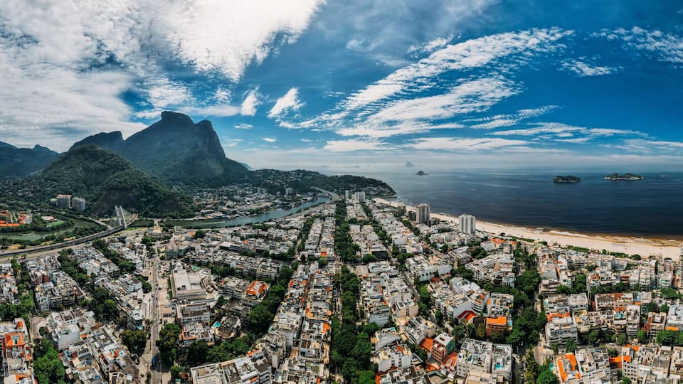 Uma vista panorâmica de uma cidade costeira com uma cadeia de montanhas imponente, uma praia e uma área urbana densa com muitos edifícios.