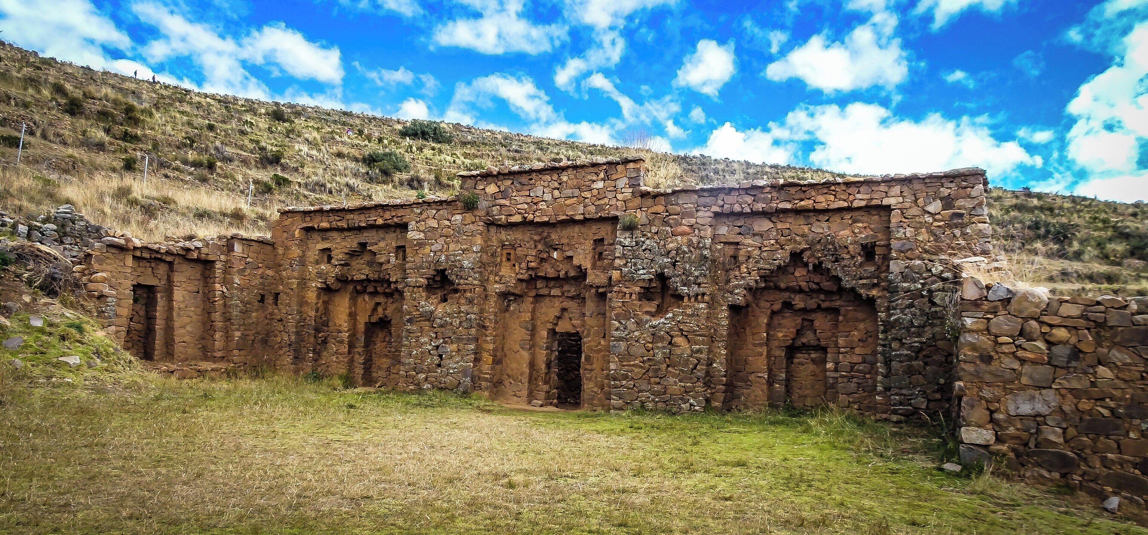 An ancient stone structure with arched doorways, set against a hillside under a partly cloudy sky.