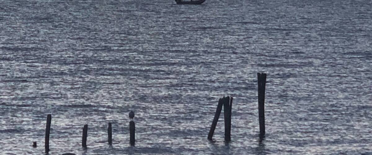 A calm sea with a boat in the distance, a mountain range in the background, and wooden posts in the foreground.