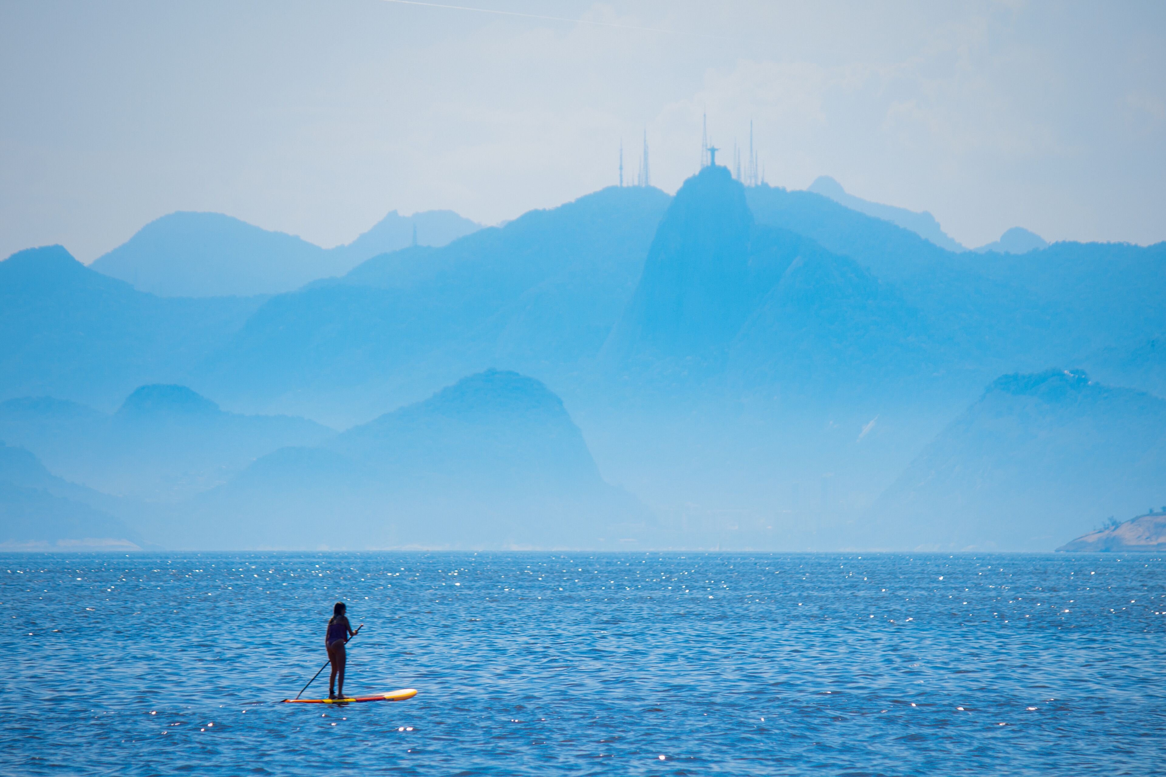 Woman stand up paddle on Camboinhas beach in Niteroi, surfing with Christ the Redeemer statue and the mountains of Rio de Janeiro in the background, clouds separating the mountains from the blue sea.