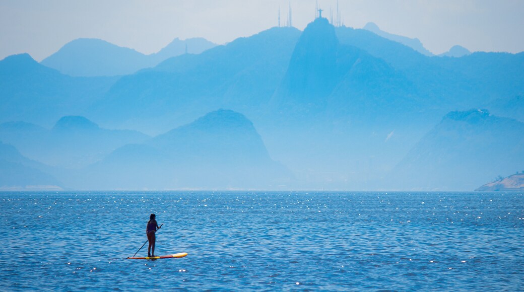 Woman stand up paddle on Camboinhas beach in Niteroi, surfing with Christ the Redeemer statue and the mountains of Rio de Janeiro in the background, clouds separating the mountains from the blue sea.