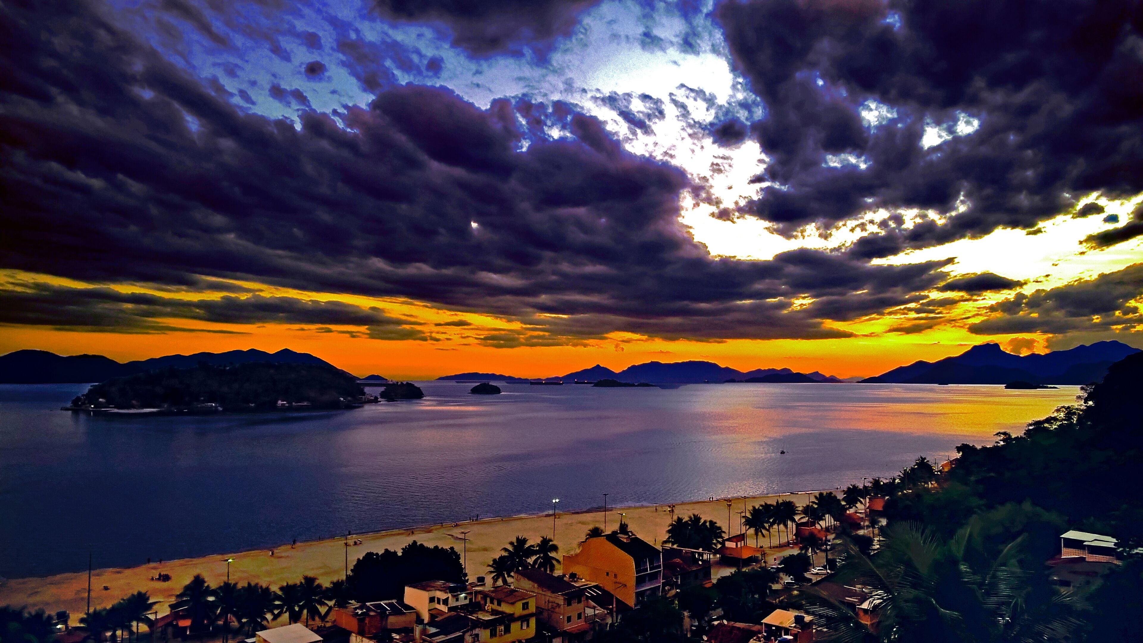 A coastal landscape with a sunset, mountains in the background, and a beach with buildings and palm trees.