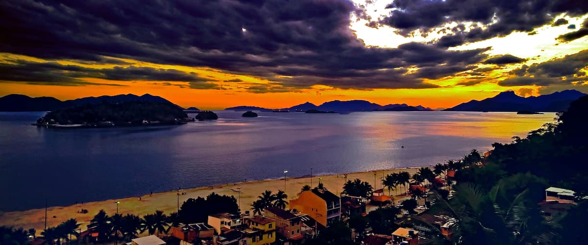 A coastal landscape with a sunset, mountains in the background, and a beach with buildings and palm trees.
