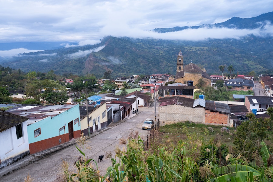 A town with a prominent church tower, a winding street, and lush green mountains in the background.