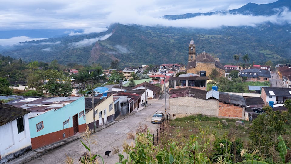 A town with a prominent church tower, a winding street, and lush green mountains in the background.