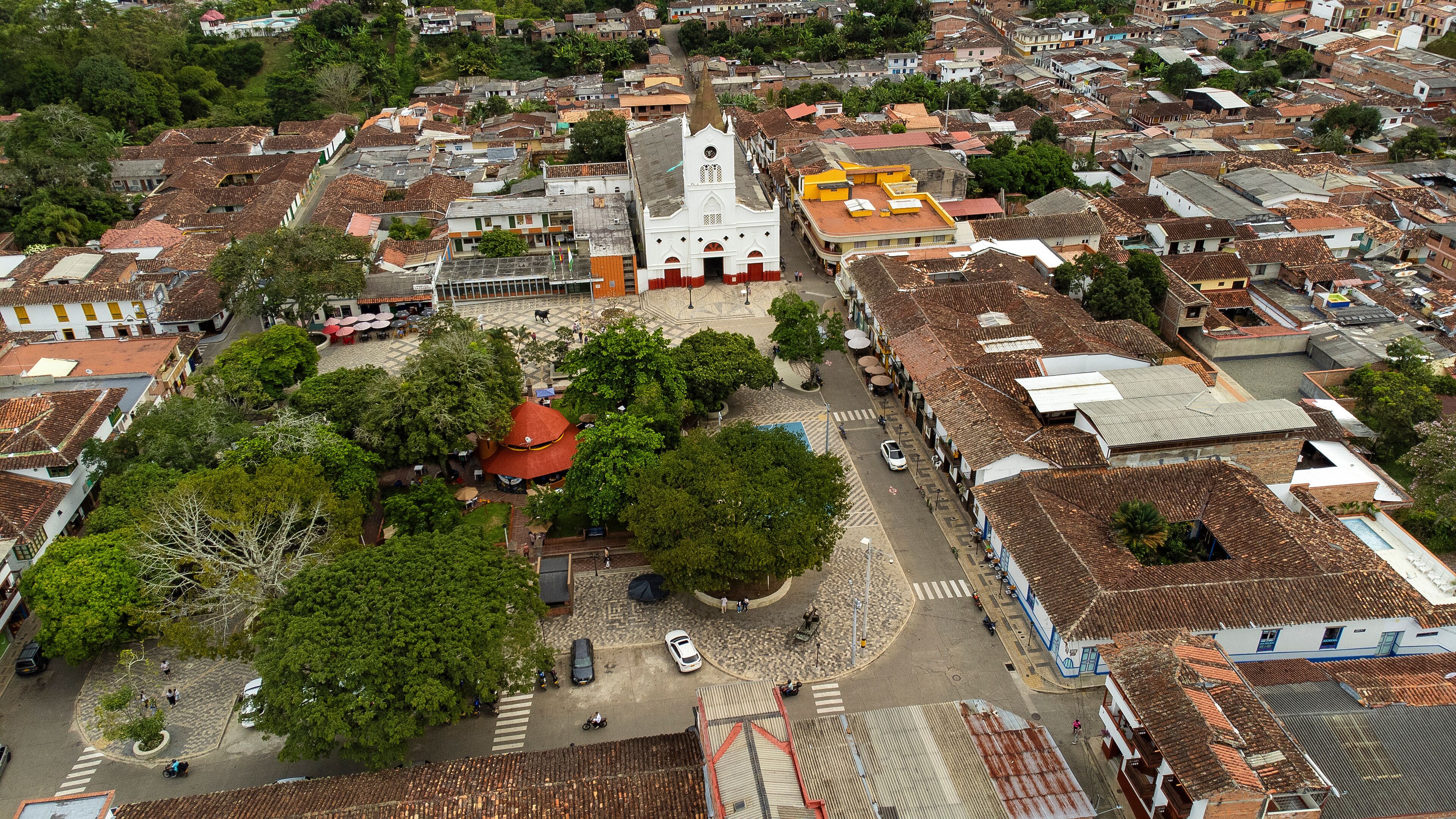 An aerial view of a town with a prominent church, a plaza with trees, and a mix of residential buildings.