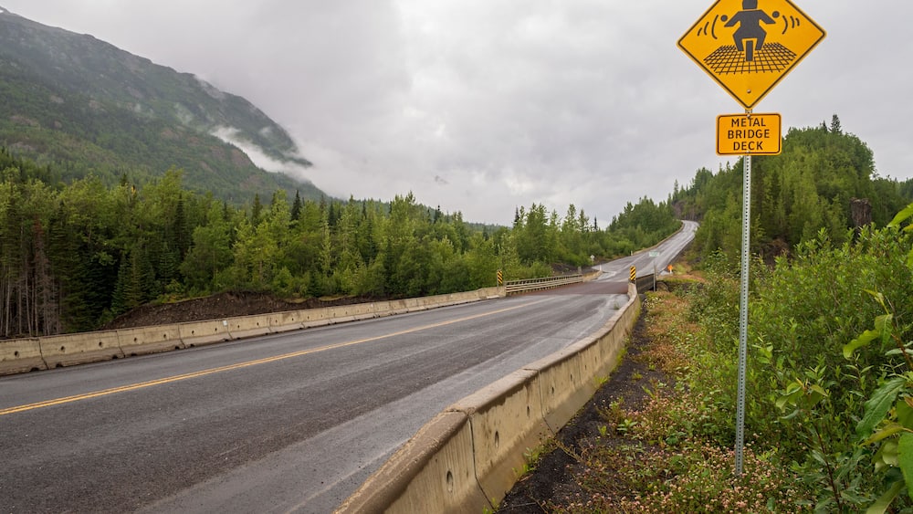 A winding road with a metal bridge deck sign and a mountain backdrop.