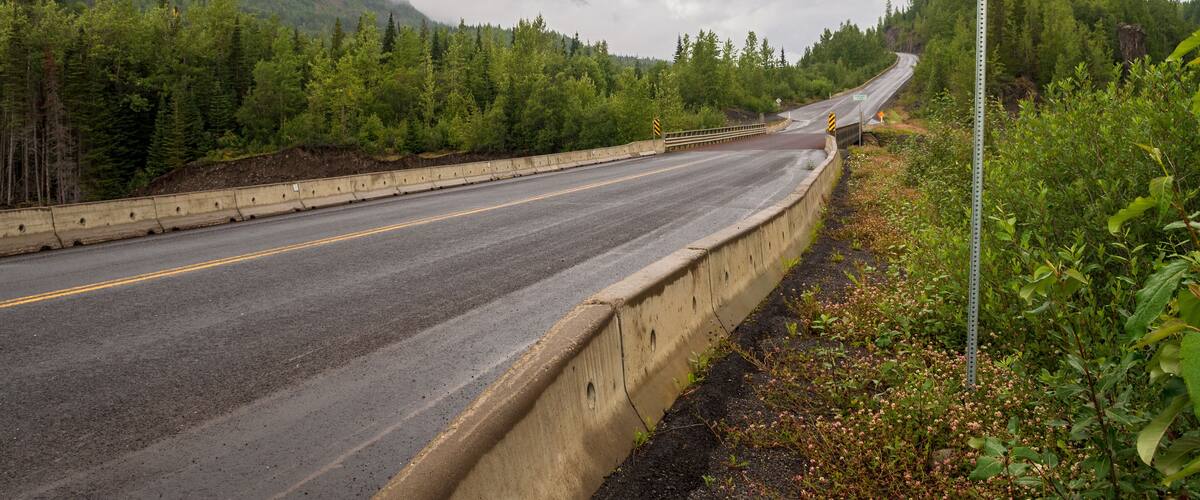 A winding road with a metal bridge deck sign and a mountain backdrop.