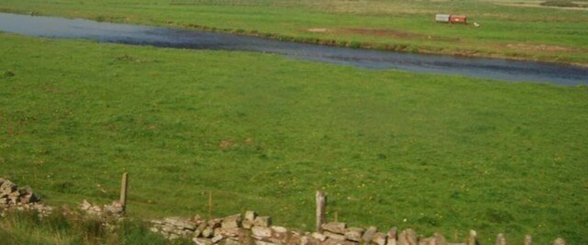 A river flowing through a green field with a stone wall in the foreground and a red vehicle in the distance.