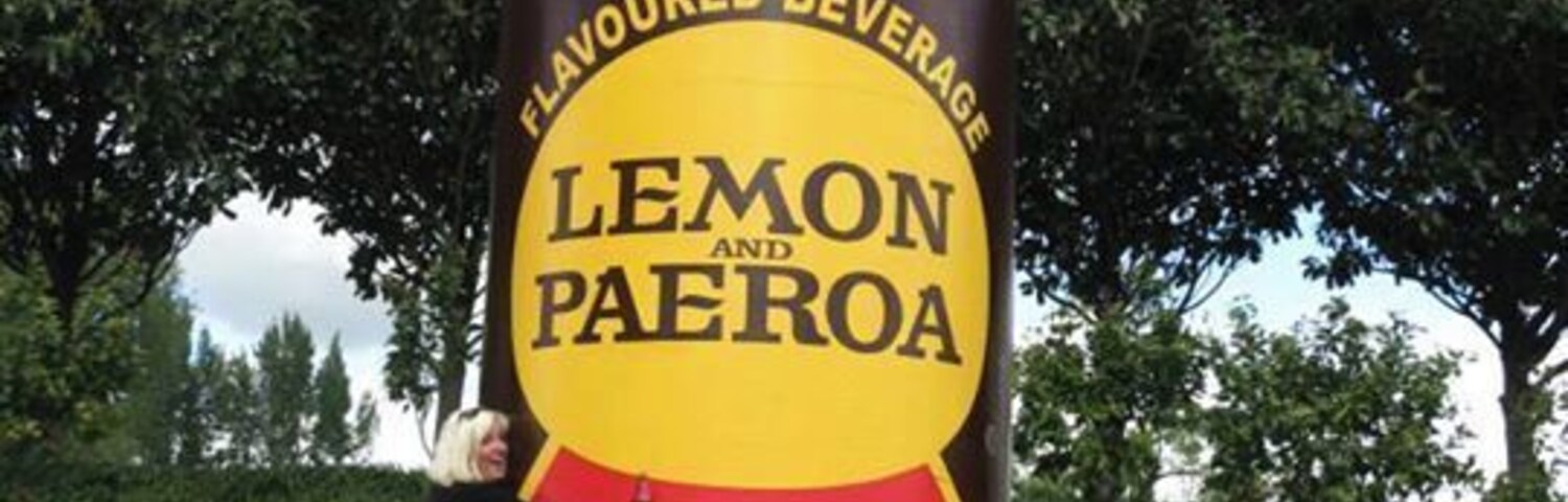 Una gran escultura de una botella con la etiqueta 'Lemon Paeroa', con una persona apoyada en ella.