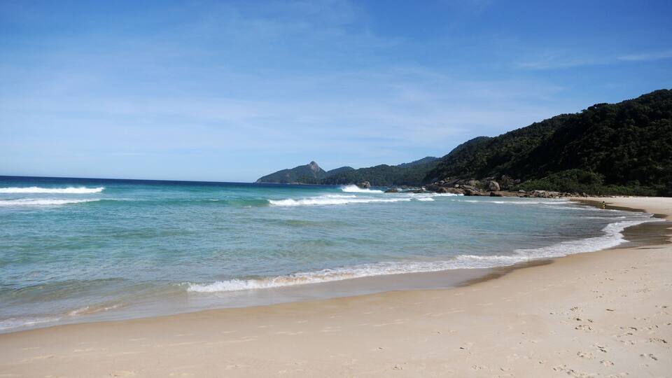 A sandy beach with clear blue water and rocky hills in the background.