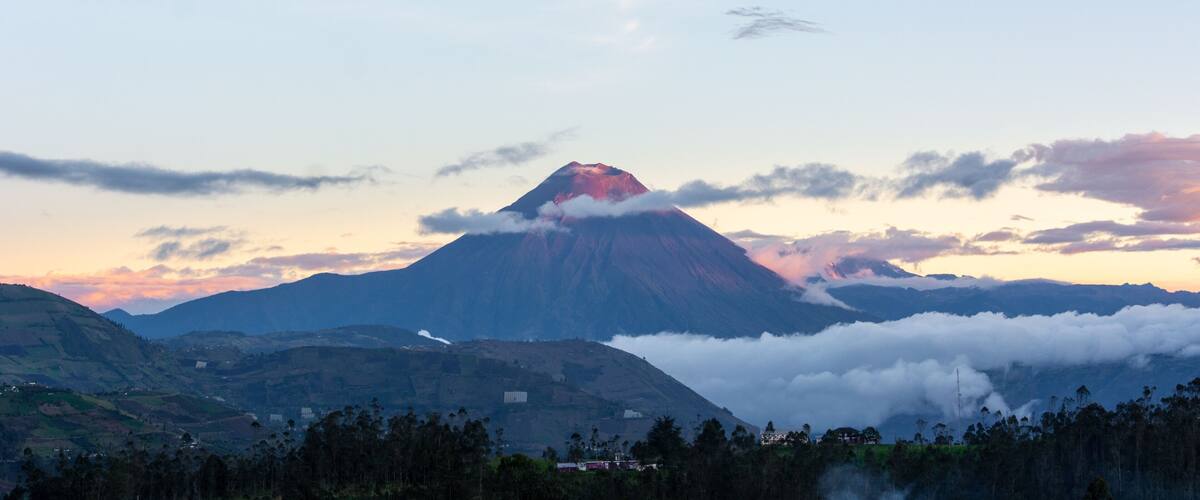 A mountain with a red glow at its peak, surrounded by clouds and forested valleys.