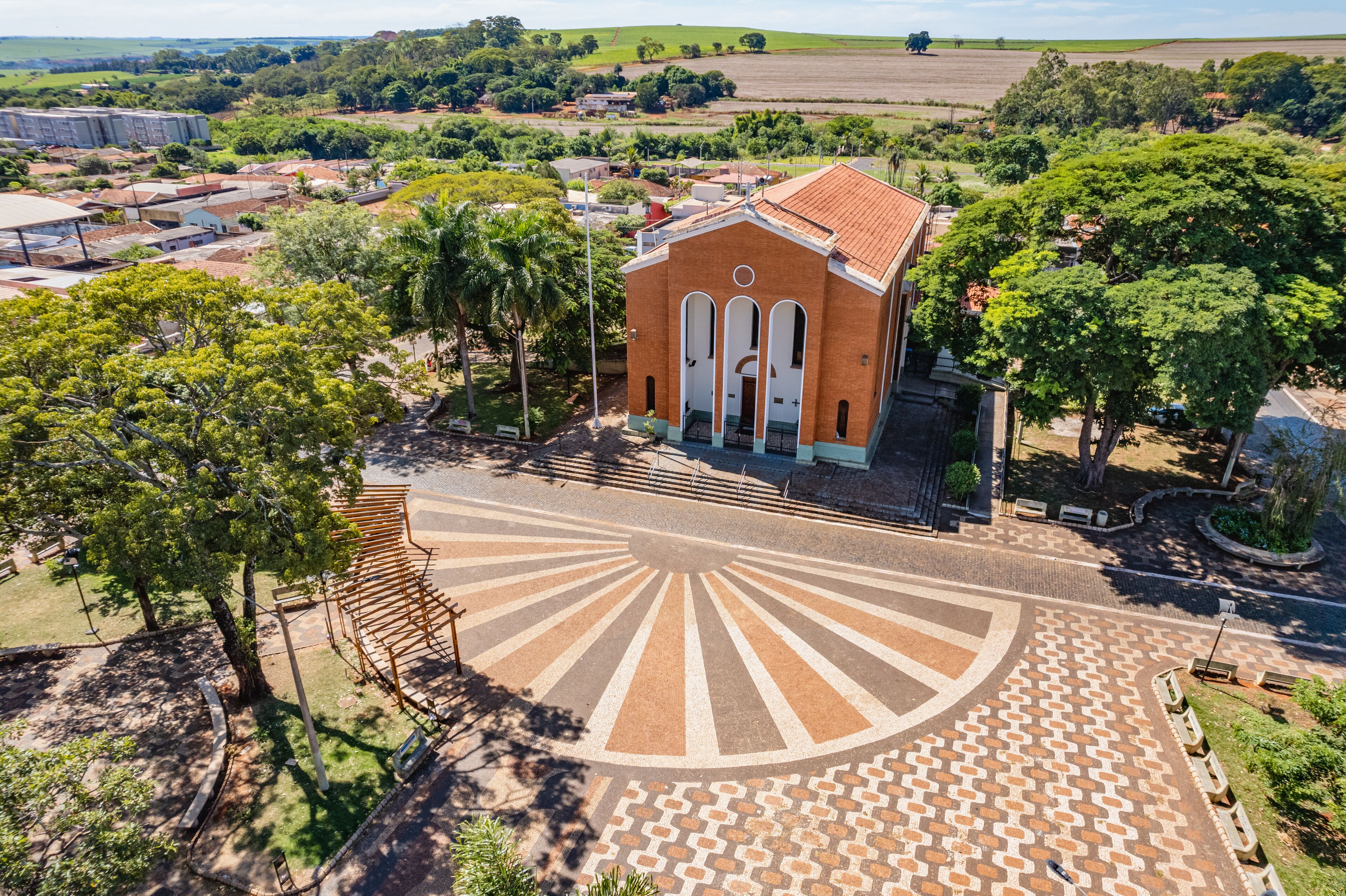 An aerial view of a church with a sunburst pavement design, surrounded by trees and fields.