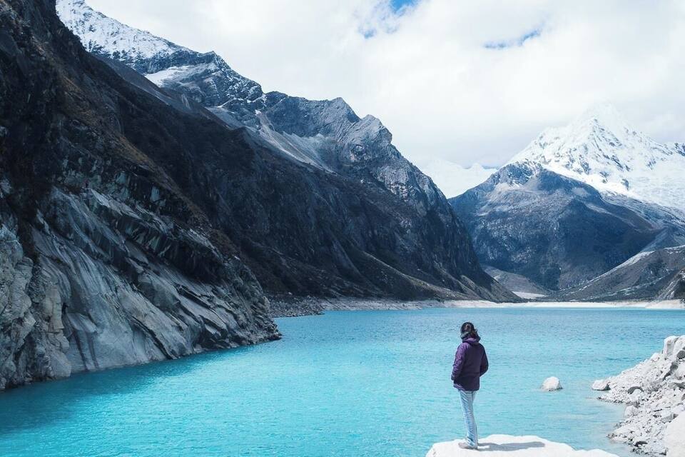 A person standing on a rock by a turquoise lake with snow-capped mountains in the background.