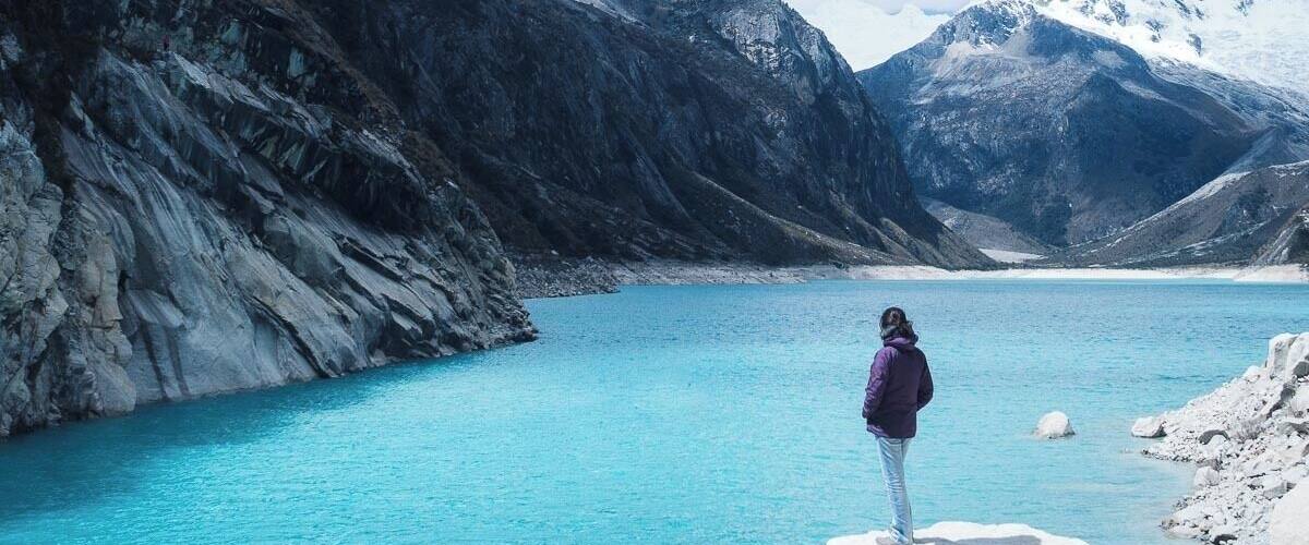 A person standing on a rock by a turquoise lake with snow-capped mountains in the background.