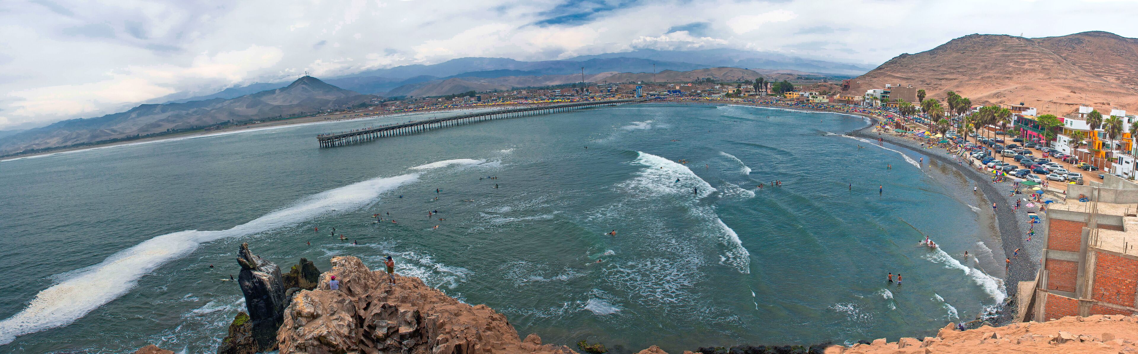 A coastal city with a long pier extending into the sea, a beach with people, and mountains in the background.