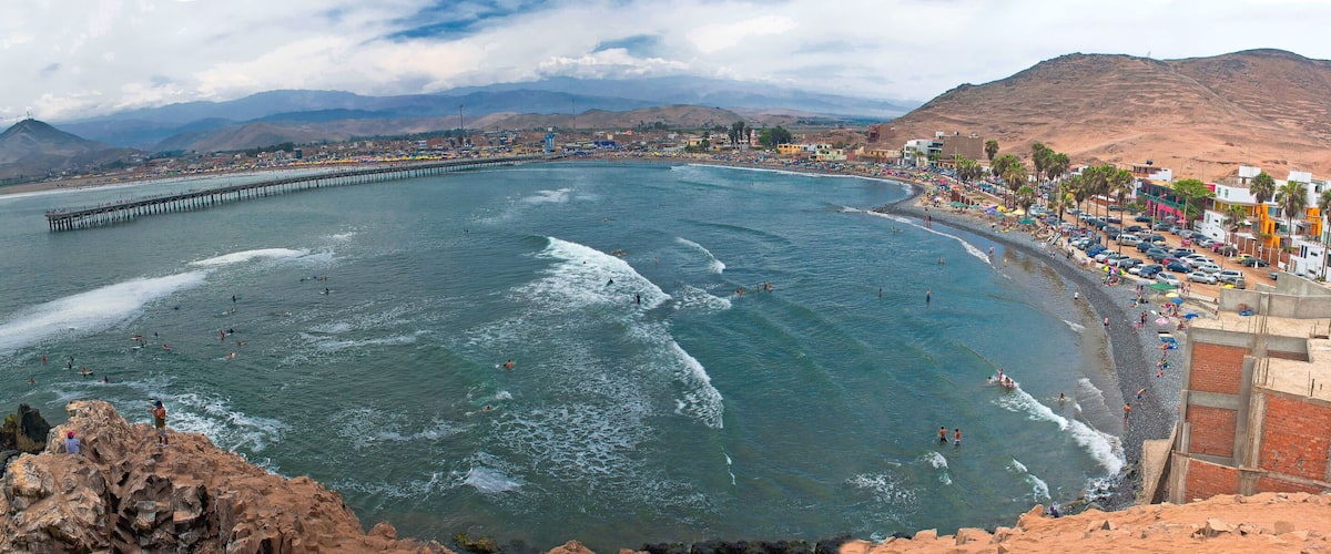 A coastal city with a long pier extending into the sea, a beach with people, and mountains in the background.