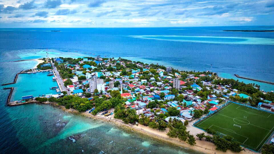 An aerial view of a coastal town with a clear blue sea, a sandy beach, and a soccer field.