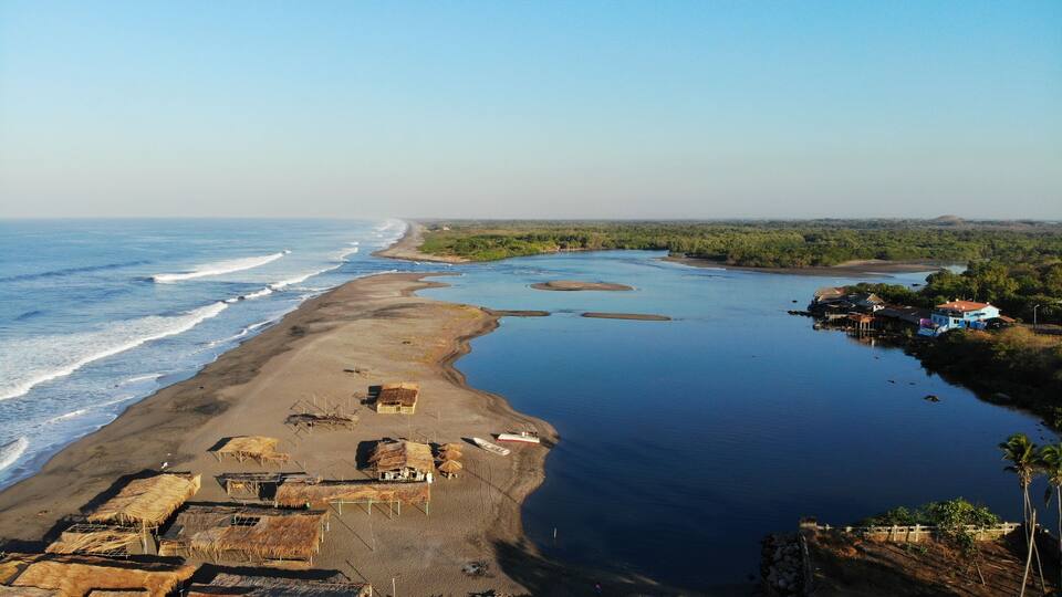 A coastal landscape with a sandy beach, a calm river, and a small settlement with houses and boats.
