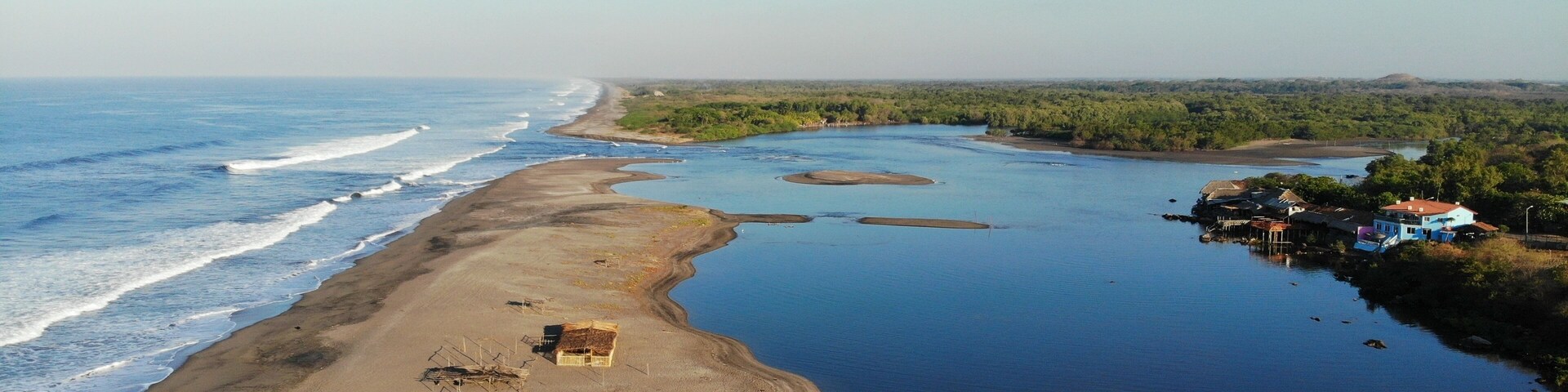 Paisagem costeira com praia arenosa, rio calmo e um pequeno povoado com casas e barcos.