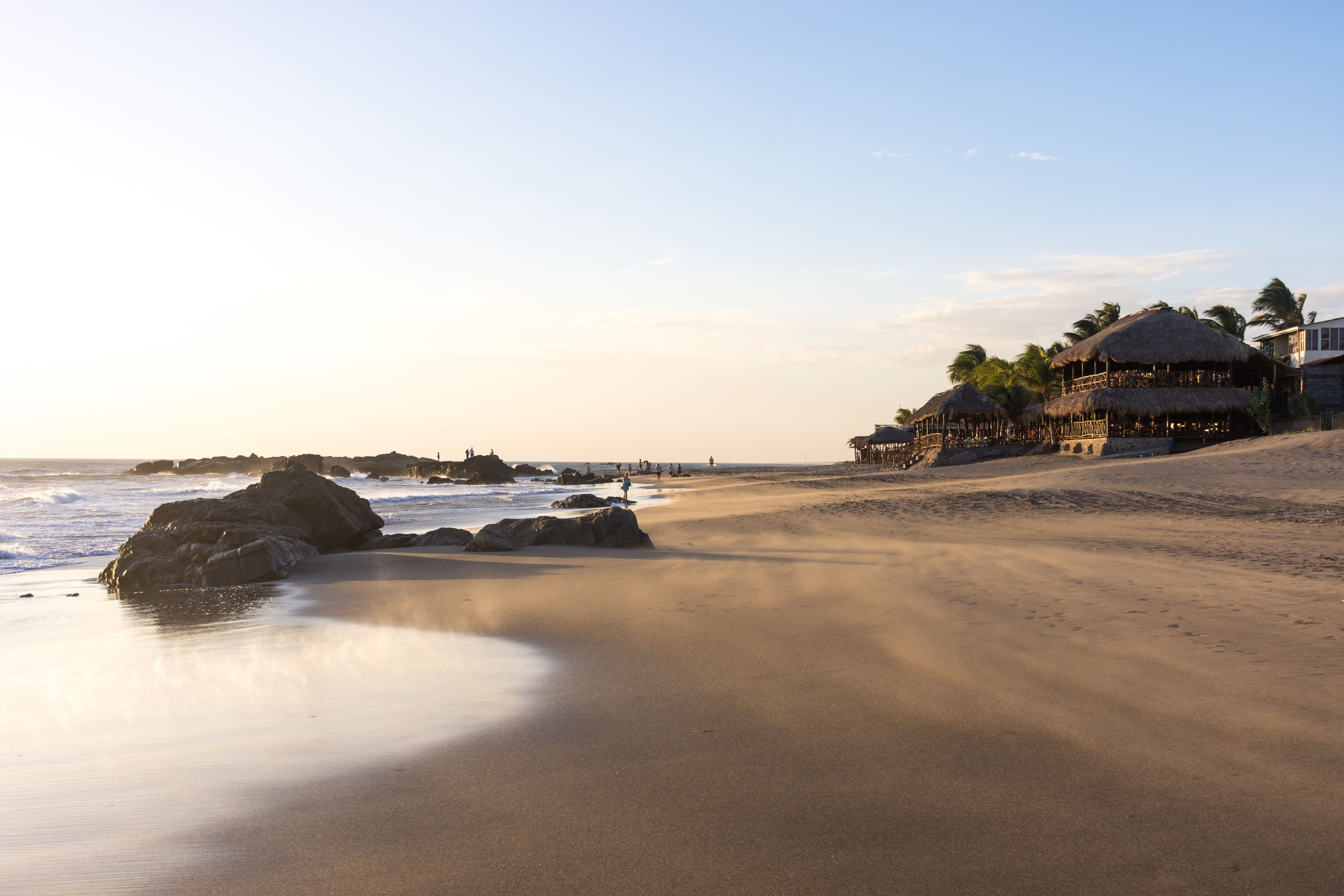 Ein Strand mit einem Gebäude mit Reetdach, Palmen und Felsen.