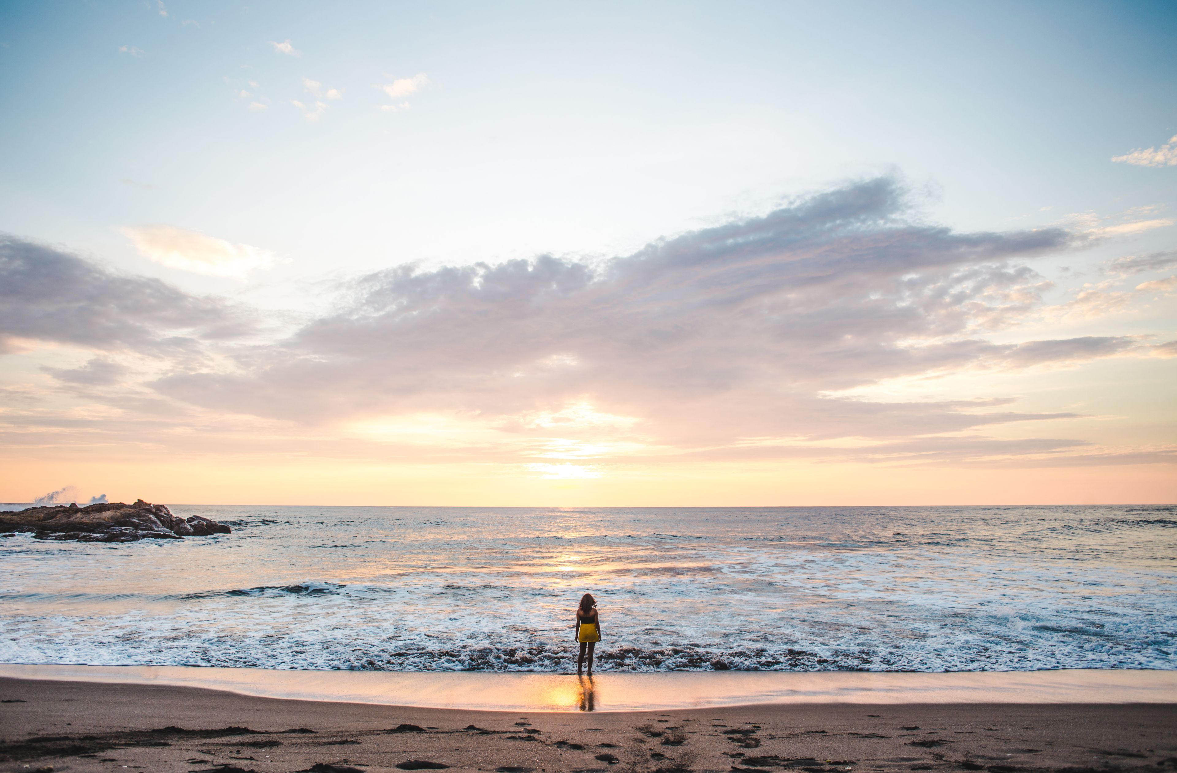 Ein Strand bei Sonnenuntergang, mit Wellen, einer Person, die am Sand steht, und einem Felsen im Hintergrund.