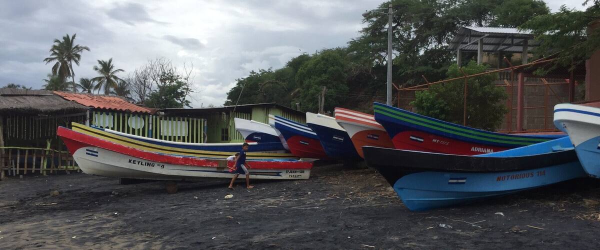 Several colorful boats parked on a sandy beach with a thatched-roof hut and palm trees in the background.