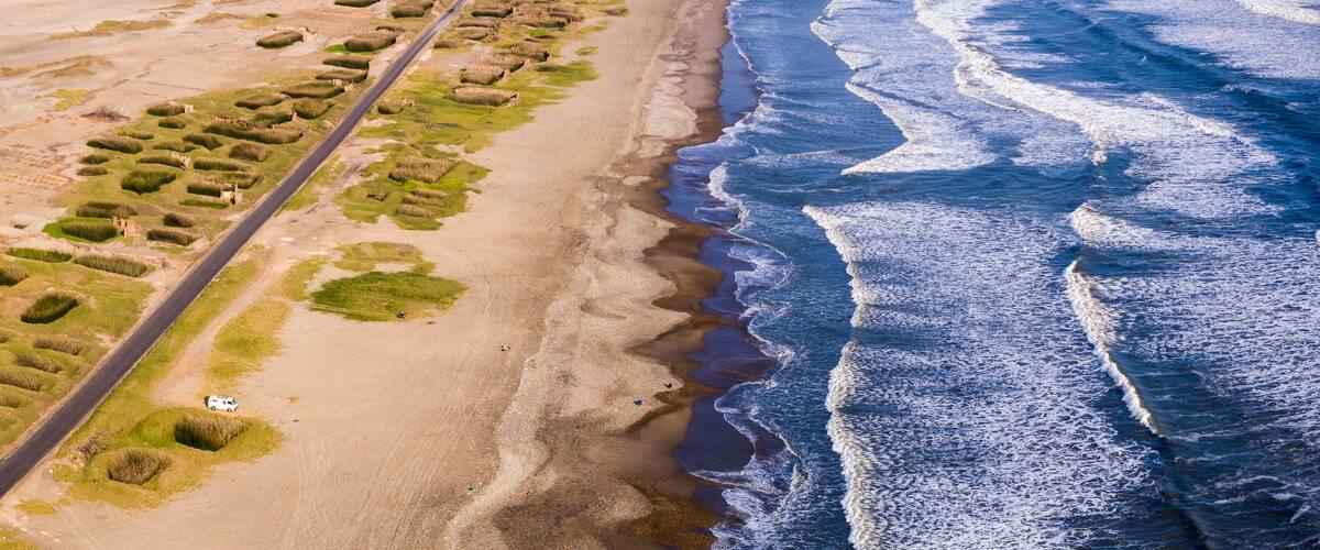 An aerial view of a coastal area with a road, grassy dunes, and ocean waves.
