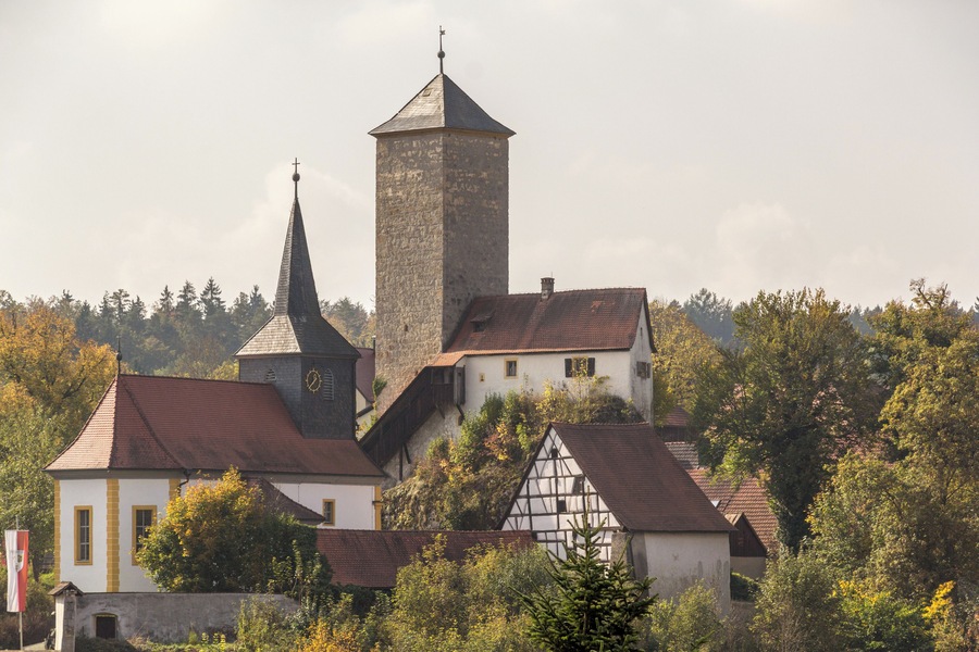 Eine Kirche mit einem hohen Turm und einem kleineren Kirchturm, umgeben von Bäumen und traditionellen Gebäuden.